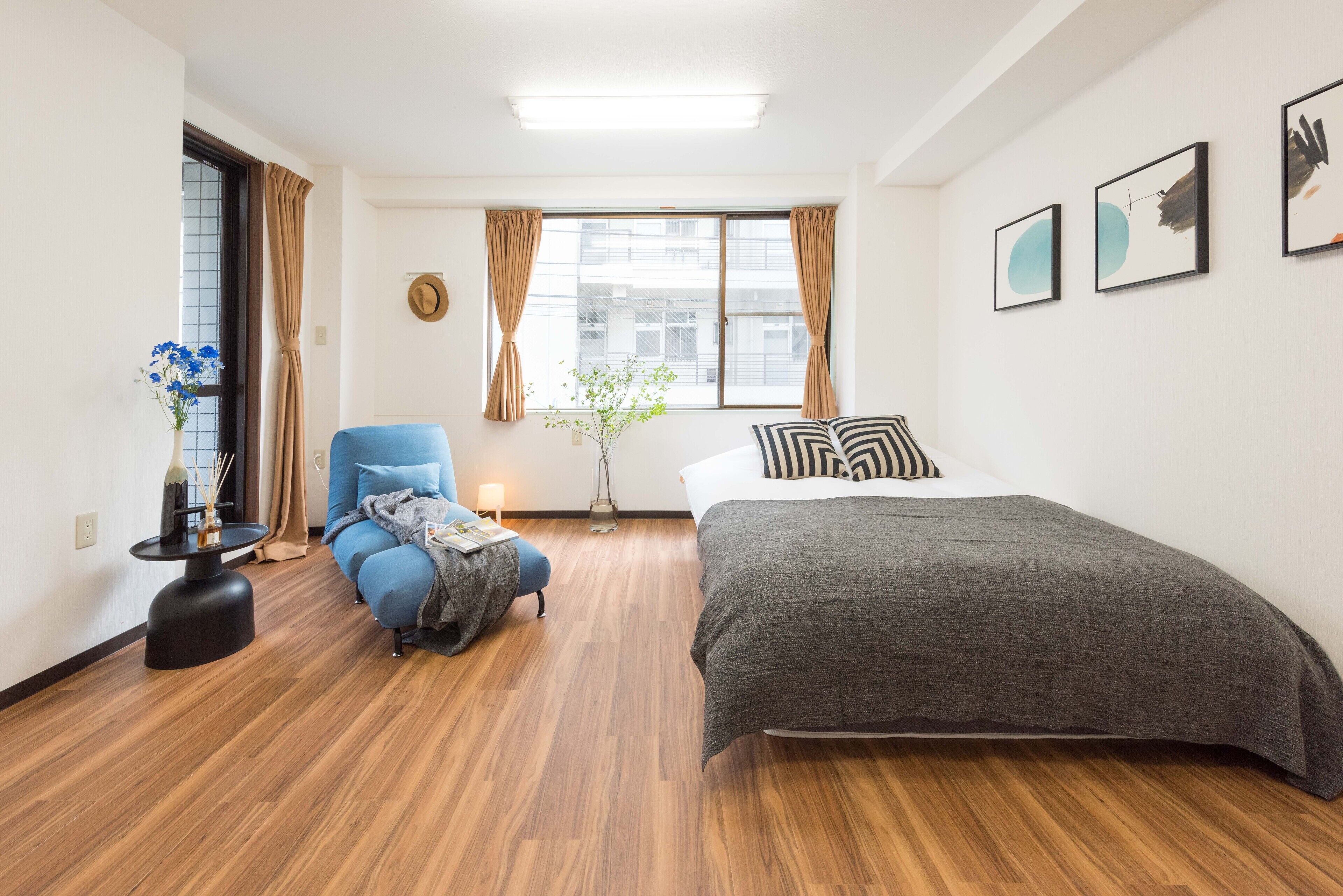 A modern bedroom with a wooden floor, a bed with a grey duvet, a blue armchair, a small table with a vase of flowers, and a window with curtains.