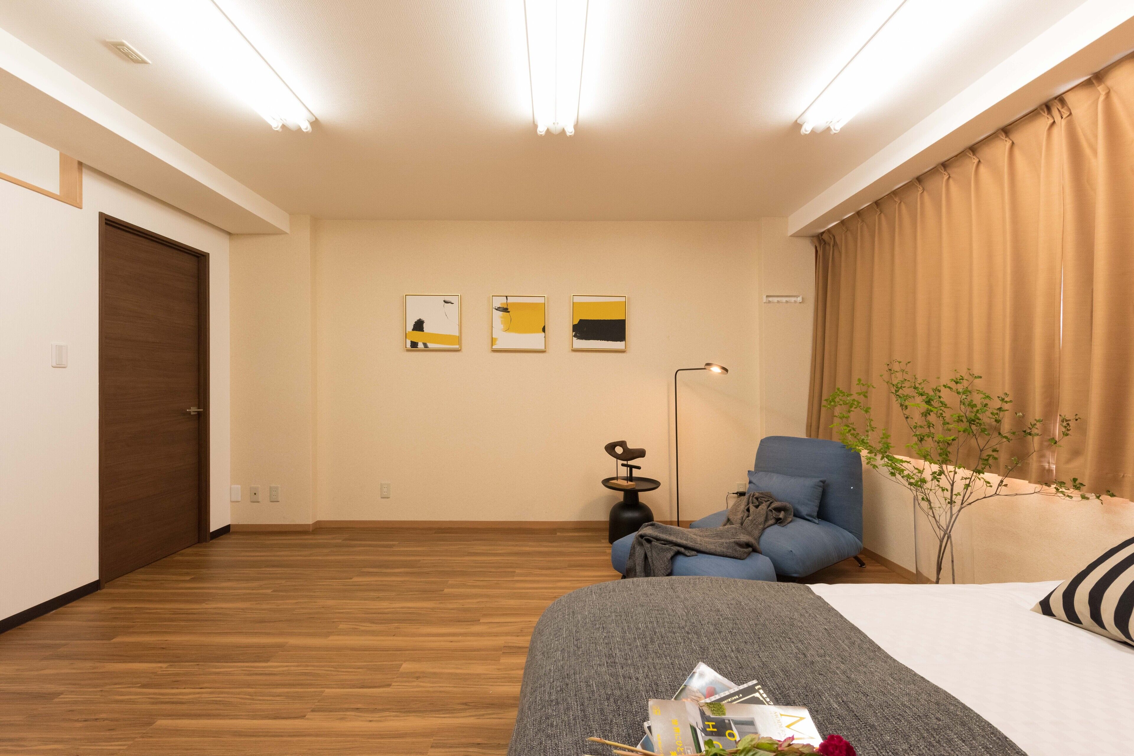 A modern hotel room with a wooden floor, a bed with a grey blanket, a blue armchair, a small side table, and three abstract wall art pieces.