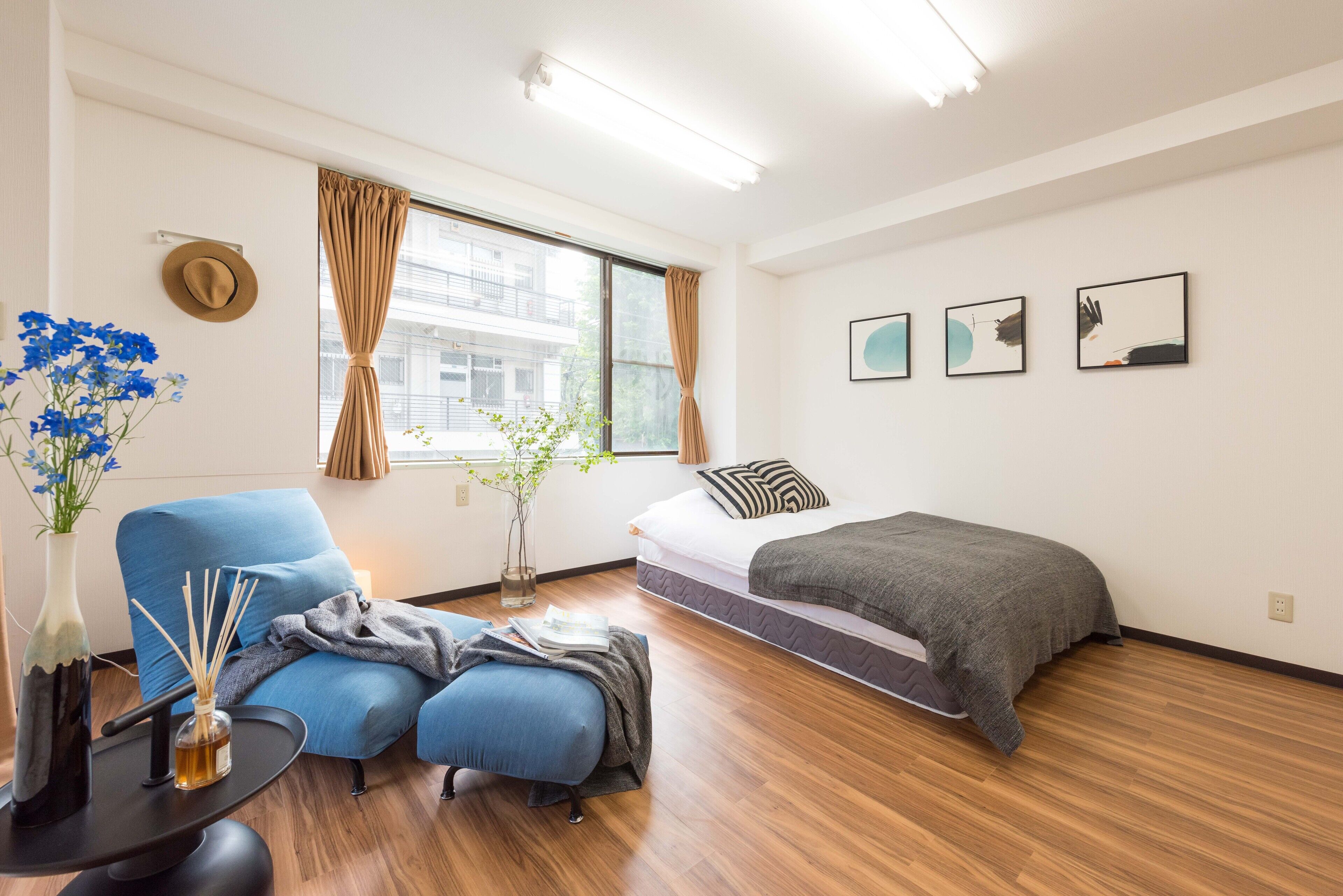 Quarto de hotel moderno com uma cama, uma poltrona azul, uma mesinha com um vaso e velas, e uma janela com cortinas.