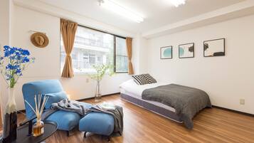 Quarto de hotel moderno com uma cama, uma poltrona azul, uma mesinha com um vaso e velas, e uma janela com cortinas.