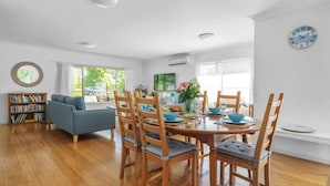 A modern living room with a dining area, wooden furniture, a bookshelf, and a large window with a view. - Elaine at Hyams Beach, Jervis Bay (Hyams Beach)
