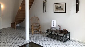 Ein Zimmer mit einem gemusterten Boden, einer Holztreppe, einem Korbstuhl, einem Glastisch mit Dekoration und Wandleuchten.