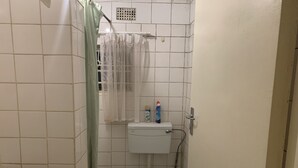A small, tiled bathroom with a toilet, a shower curtain, and a shelf with toiletries. - Furnished Self Catering cottage (Lusaka)