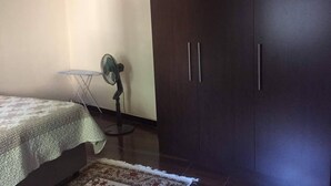 A bedroom with a bed, a dark wardrobe, a standing fan, and a small table. - Furnished Self Catering cottage (Lusaka)