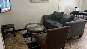 A living room with a grey sofa, a brown armchair, a glass-top coffee table, a small black side table, and a TV mounted on the wall. - Furnished Self Catering cottage (Lusaka)
