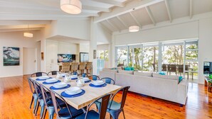 A spacious dining area with a long wooden table set for a meal, a large window with a view of the outdoors, and a cozy seating area with cushions.