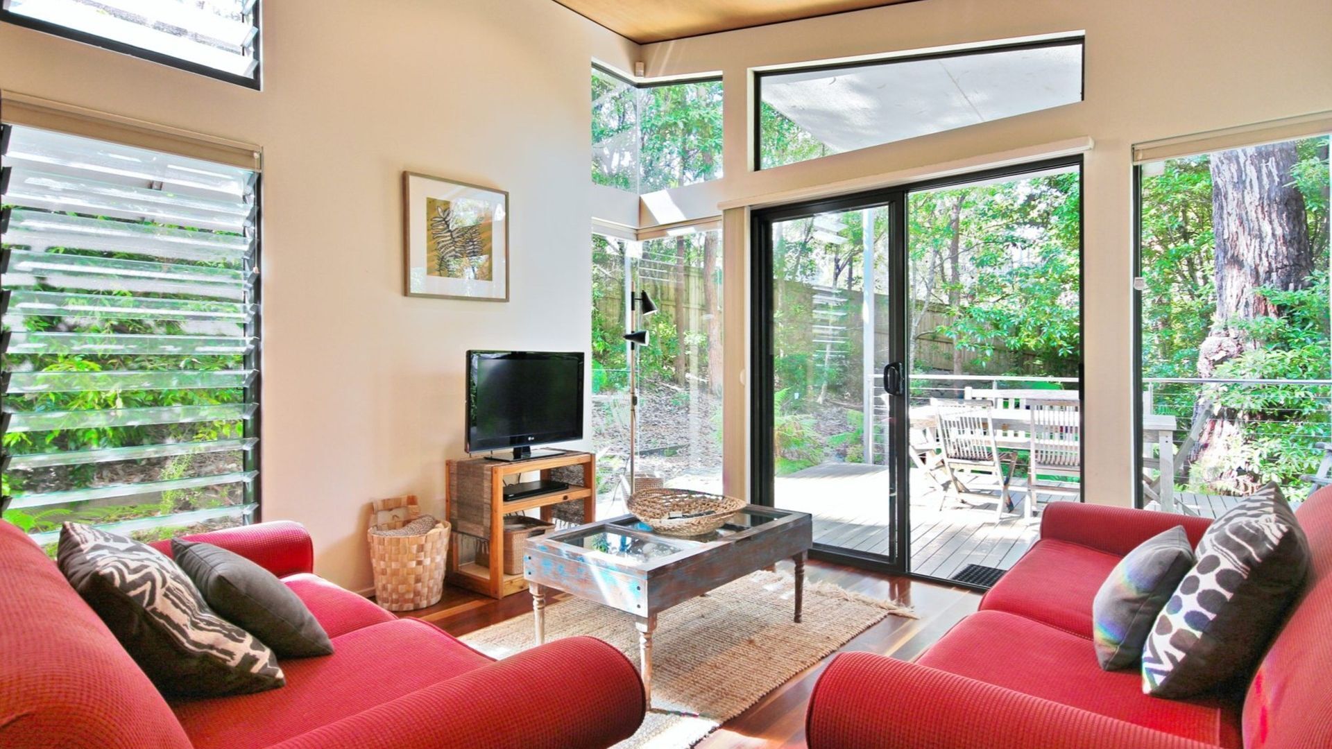 A modern living room with a large TV, red sofas, a glass coffee table, and a view of a deck and trees.