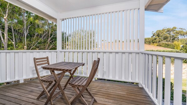 A covered outdoor area with a wooden table and two chairs, surrounded by white railings and a view of trees and a building.