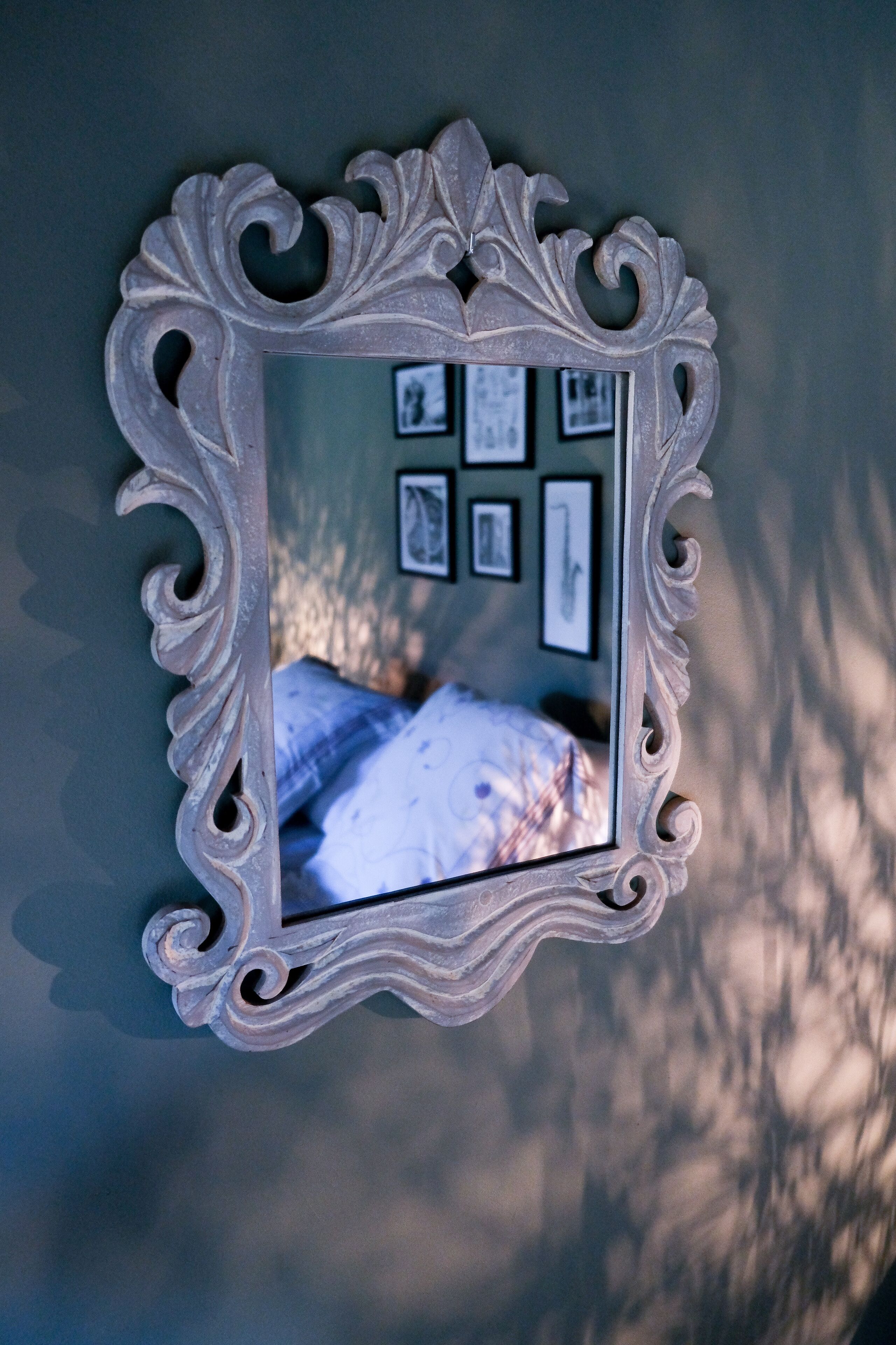 Uno specchio con una cornice decorata riflette un letto e una parete con quadri appesi.