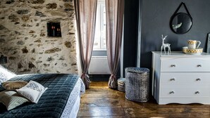 A bedroom with a stone wall, wooden floor, a bed with a quilt, a dresser, a wicker basket, and a mirror on the wall. - Au nid de Caroline (ISSOR)