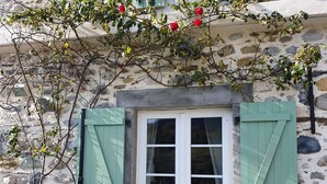 A stone building with a white door and green shutters, surrounded by gravel and a flowering vine. - Au nid de Caroline (ISSOR)