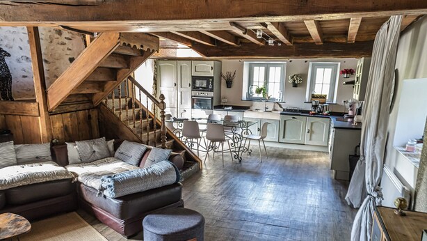 A rustic interior with exposed wooden beams, a cozy seating area with a sofa and ottoman, a kitchen with a central island, and a staircase leading to an upper level. - Au nid de Caroline (ISSOR)
