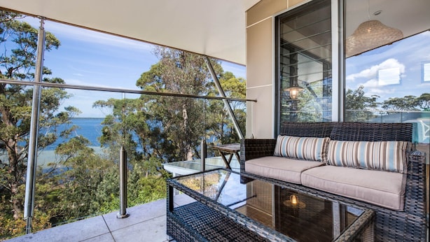 A balcony with a glass balustrade, a wicker sofa with striped cushions, and a view of the sea and trees. - Majestic on Palm Beach, Jervis Bay (Sanctuary Point)