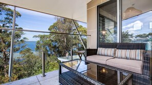 A balcony with a glass balustrade, a wicker sofa with striped cushions, and a view of the sea and trees. - Majestic on Palm Beach, Jervis Bay (Sanctuary Point)