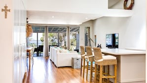 A modern living room with a sofa, chairs, a TV, and a view of the outdoors. - Majestic on Palm Beach, Jervis Bay (Sanctuary Point)