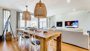 A modern living room with a wooden dining table, white chairs, a white sofa, and a television mounted on the wall. - Majestic on Palm Beach, Jervis Bay (Sanctuary Point)