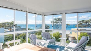 A sunlit room with large windows, wicker chairs, a wooden table, and a view of the sea. - Mara at Hyams Beach, Jervis Bay (Hyams Beach)