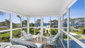 A sunroom with large windows, wicker furniture, and a view of the ocean and hills. - Mara at Hyams Beach, Jervis Bay (Hyams Beach)