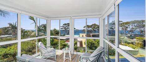A sunroom with large windows, wicker furniture, and a view of the ocean and hills.