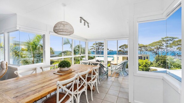 A spacious dining area with a wooden table and white chairs, large windows offering a view of the ocean and greenery, and a hanging light fixture. - Mara at Hyams Beach, Jervis Bay (Hyams Beach)