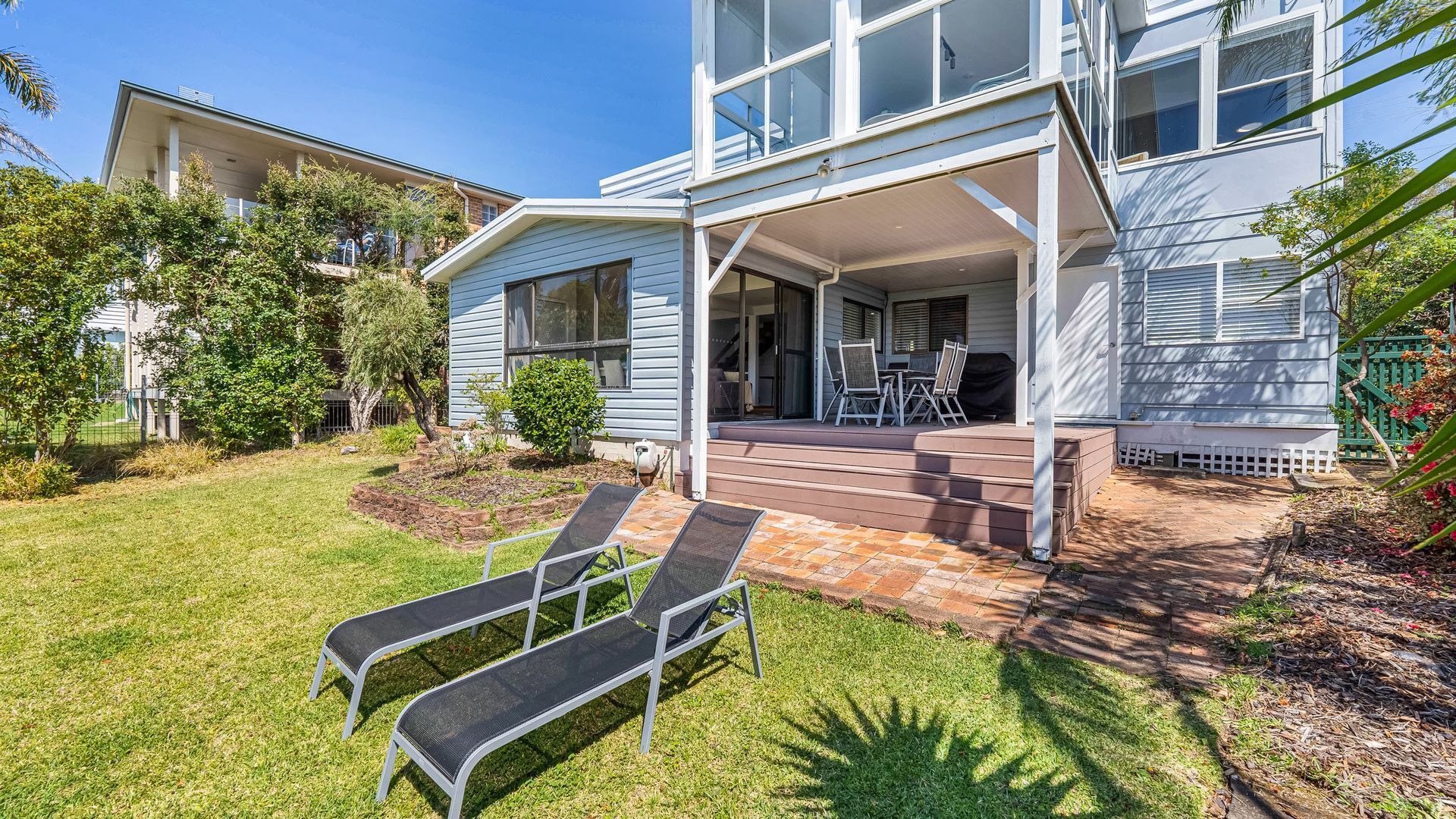 A two-story house with a covered porch, a small garden, and two lounge chairs.