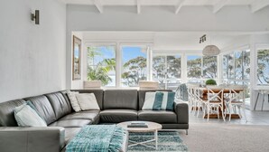 A modern living room with a leather sectional sofa, a round coffee table, and a dining area with white chairs and a wooden table. - Mara at Hyams Beach, Jervis Bay (Hyams Beach)