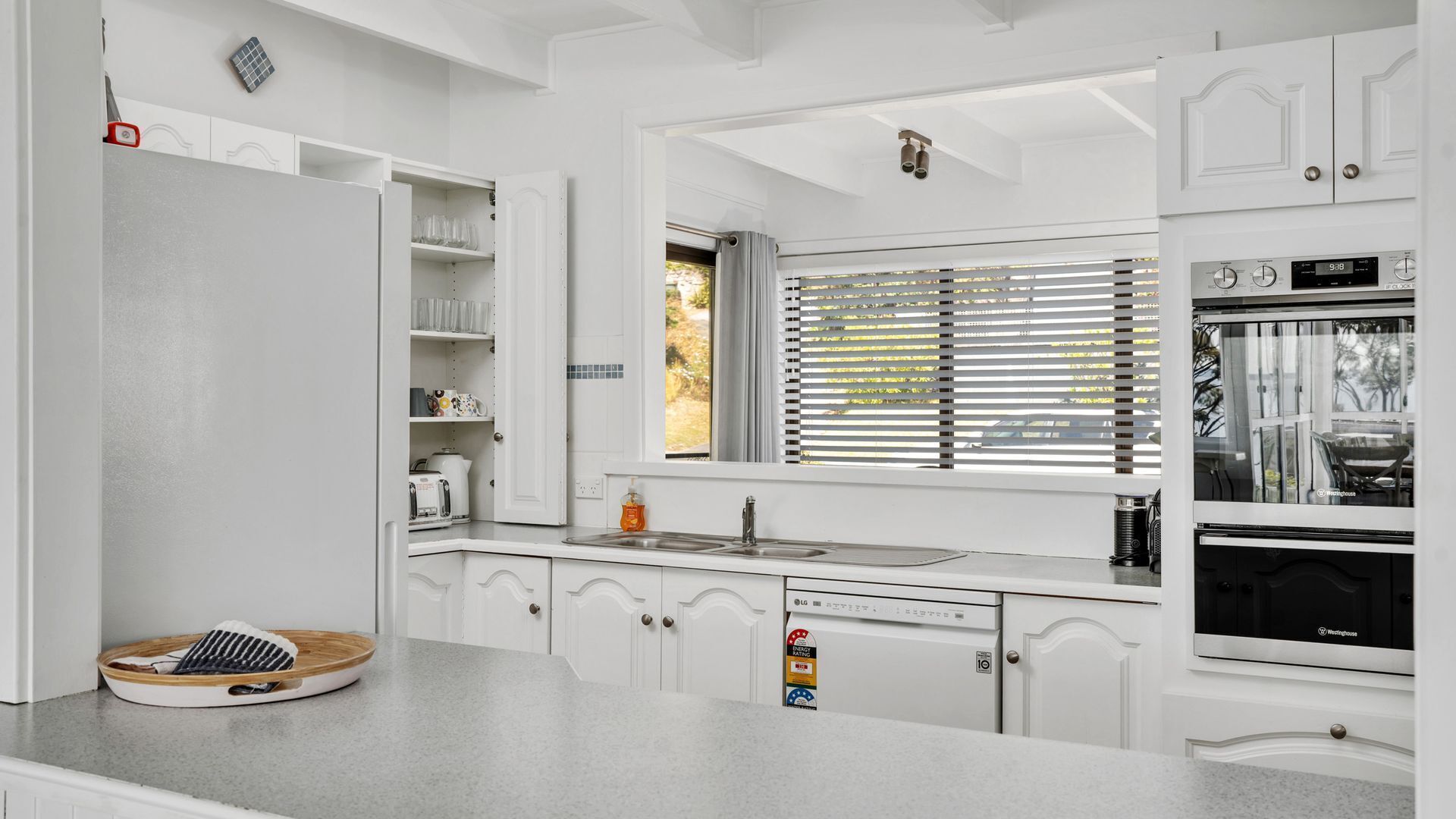 Une cuisine moderne avec des meubles blancs, un four encastré et une fenêtre équipée de stores.