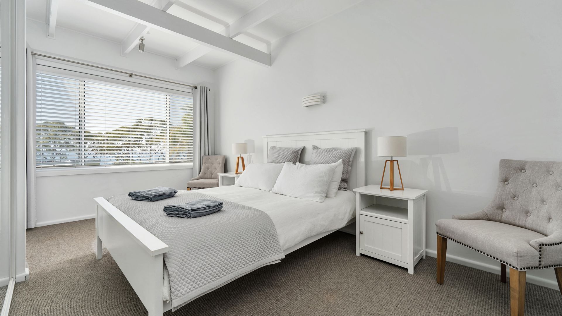 Une chambre moderne avec un lit blanc, des tables de chevet, une chaise et une fenêtre avec des stores.