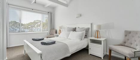 Une chambre moderne avec un lit blanc, des tables de chevet, une chaise et une fenêtre avec des stores.