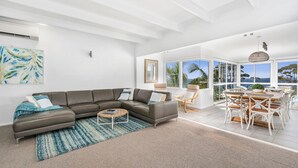 A spacious living room with a large sectional sofa, a coffee table, and a dining area with white chairs and a wooden table. - Mara at Hyams Beach, Jervis Bay (Hyams Beach)