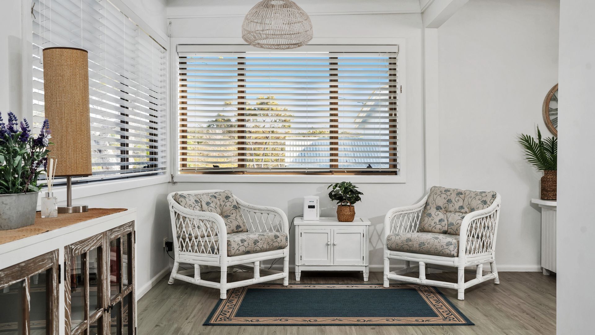 Una habitación con dos sillones blancos de mimbre, un armario pequeño blanco y un ventanal con persianas.