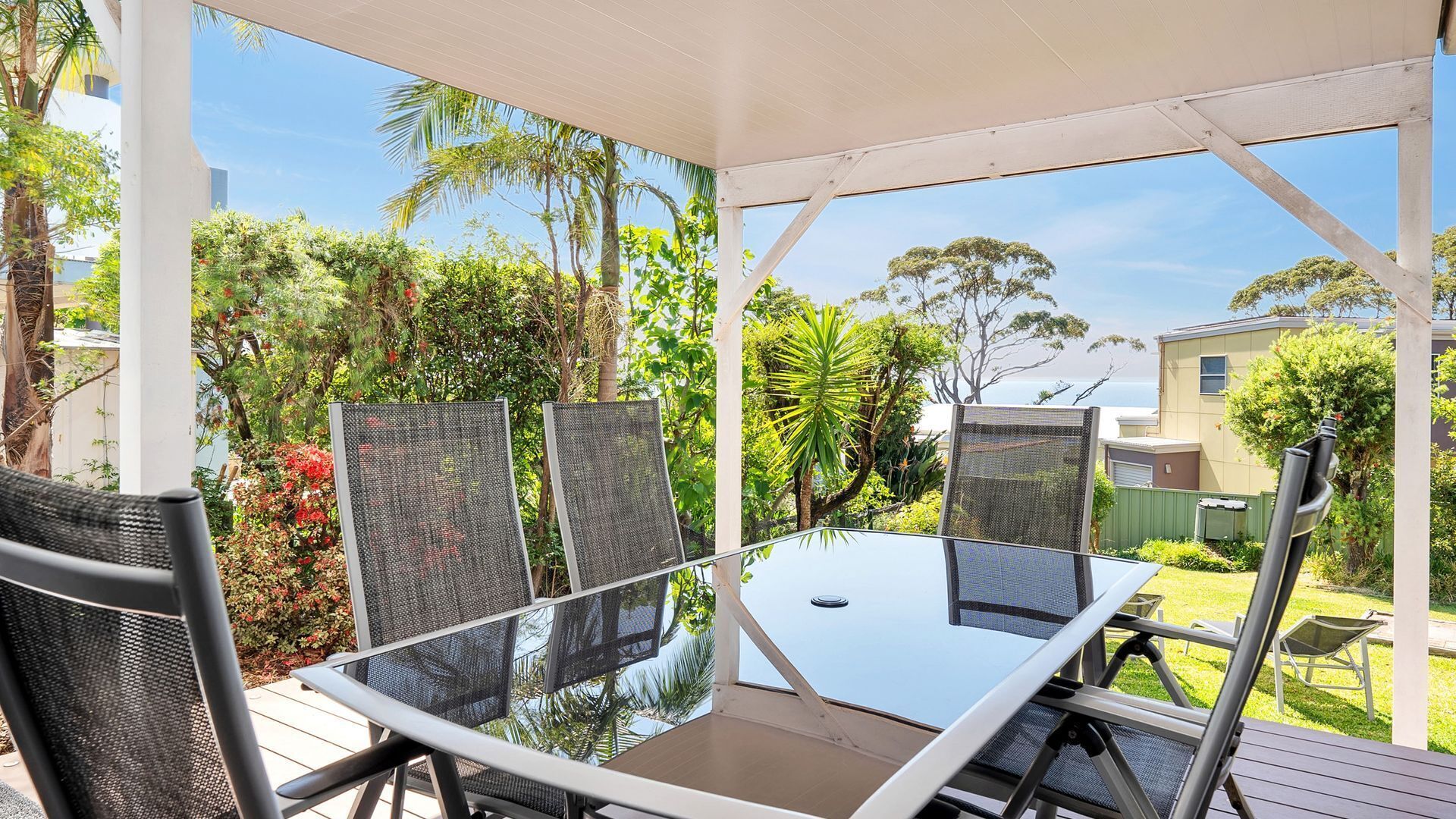 An outdoor patio with a glass table and metal chairs, surrounded by lush greenery and a view of the ocean.
