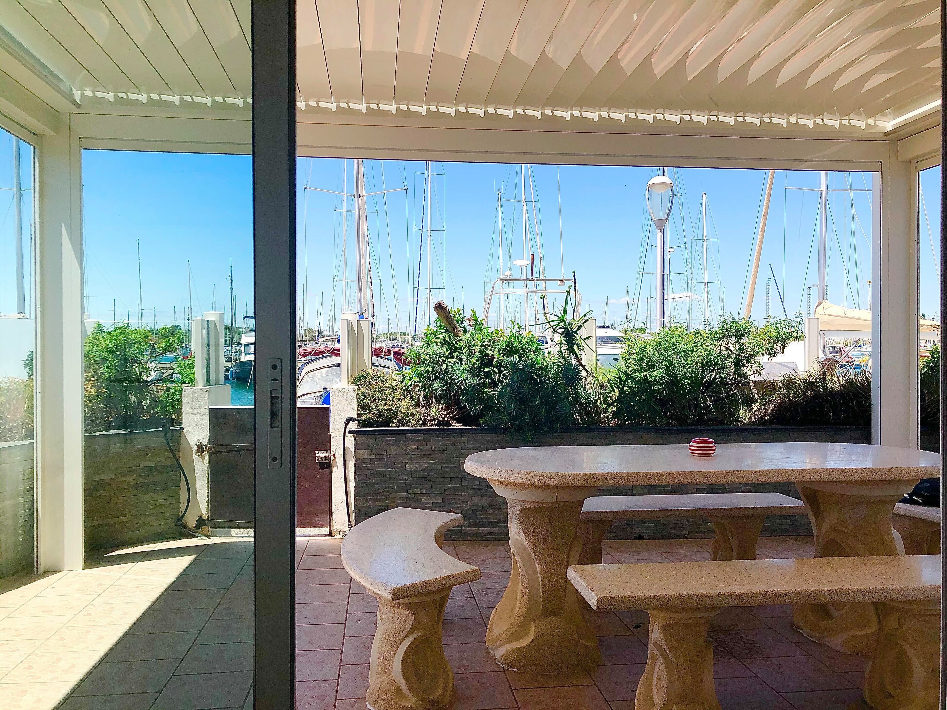 Una veranda riparata con tavolo e panche, affacciata su un porto turistico con vele.