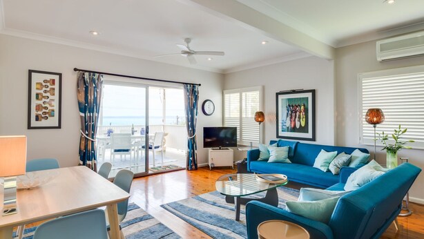 Un salon moderne avec un canapé bleu, une table à manger et un téléviseur. Il y a un balcon avec vue sur la mer.