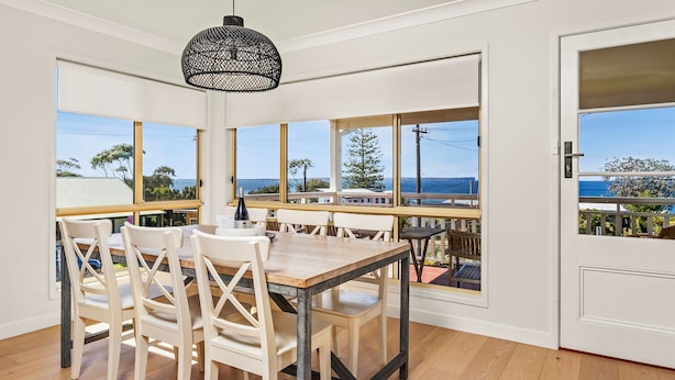 Un comedor con mesa y sillas de madera, ventanas amplias que ofrecen vistas al océano y a los árboles, y una lámpara colgante.