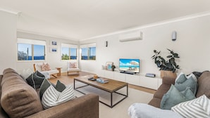 Una sala de estar moderna con un sofá, una mesa de centro y una televisión. Amplias ventanas ofrecen vistas al exterior.
