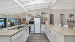 A modern kitchen with white cabinetry, a built-in oven, and a refrigerator. There is a dining area with white chairs and a view of the outdoors.