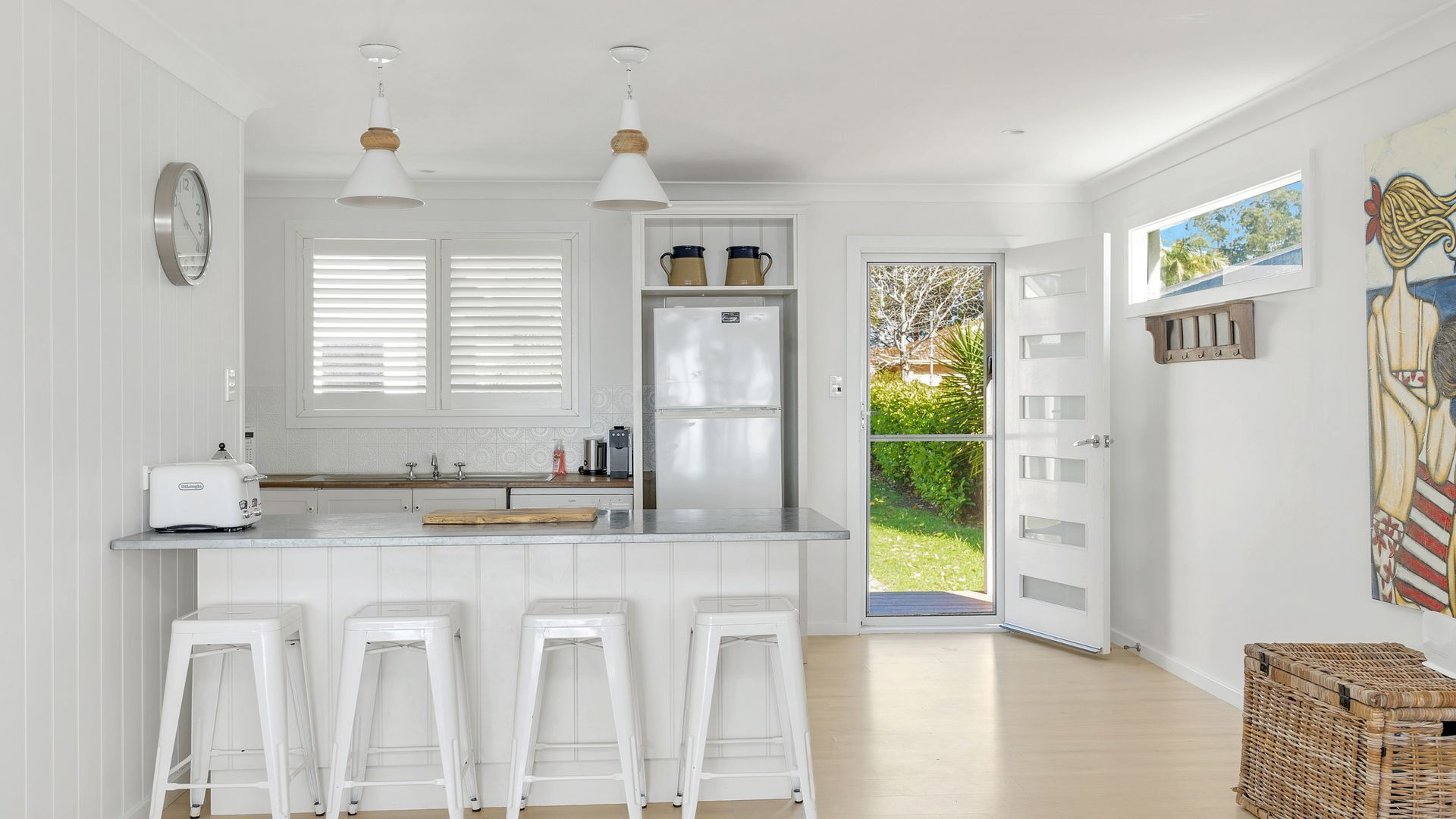 A modern kitchen with white cabinetry, a double sink, and a built-in refrigerator. There are two pendant lights, a clock on the wall, and a view of the outdoors through a glass door.