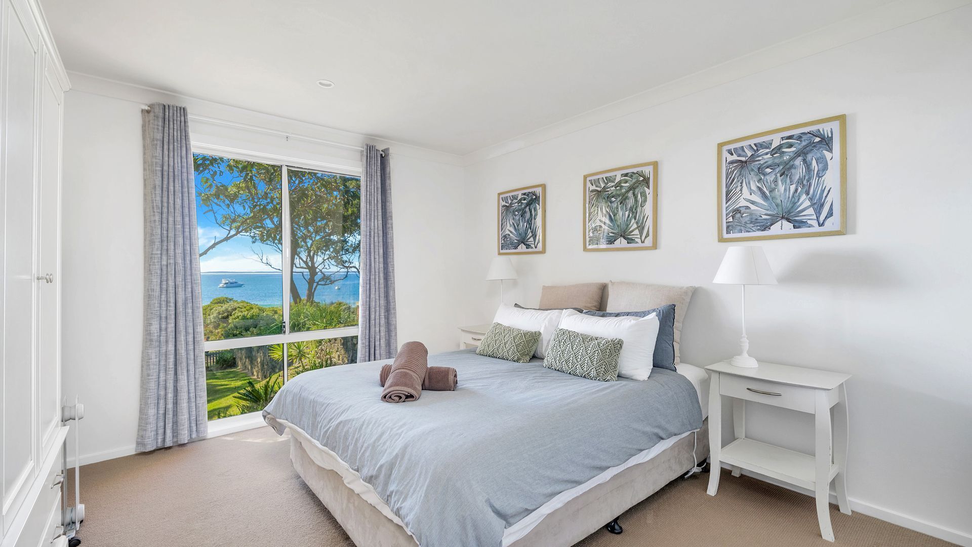 Ein Schlafzimmer mit großem Fenster und Blick auf das Meer und die grüne Landschaft. An der Wand hängen drei gerahmte Pflanzenbilder.