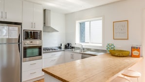 A modern kitchen with white cabinetry, stainless steel appliances, a wooden countertop, and a window with blinds. - Palmwood at Hyams Beach (Hyams Beach)