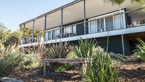 A modern two-story house with a balcony, surrounded by various plants and a wooden bench. - Palmwood at Hyams Beach (Hyams Beach)