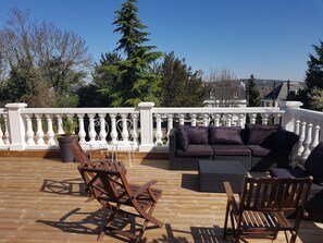Een houten terras met buitenmeubilair, zoals een bank en stoelen, en een witte balustrade.