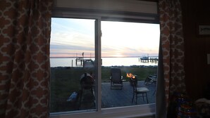 Blick durch ein Fenster auf einen Grill und Stühle draußen, mit Blick auf ein Gewässer und einen Steg.
