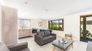 A modern living space with a kitchen, a grey sofa, a glass coffee table, and a view of greenery outside. - Sandals at the Bay - Downstairs, Hyams Beach (Hyams Beach)