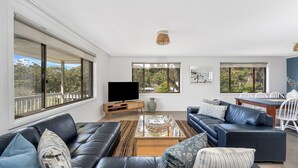 A spacious living room with a large window, a flat-screen TV, a dining area with a table and chairs, and a blue leather sofa. - Sandals at the Bay - Upstairs sleeps 6, Hyams Beach (Hyams Beach)