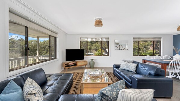 A spacious living room with a large window, a flat-screen TV, a dining area with a table and chairs, and a blue leather sofa.