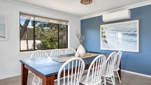 A dining area with a blue table, white chairs, a vase with dried flowers, and a framed picture of a beach scene. - Sandals at the Bay - Upstairs sleeps 6, Hyams Beach (Hyams Beach)