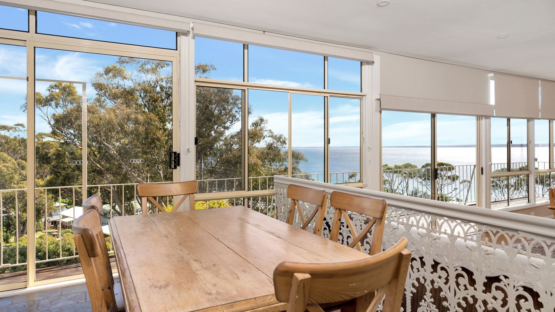 A dining area with a wooden table and chairs, large windows offering a view of the sea and trees.