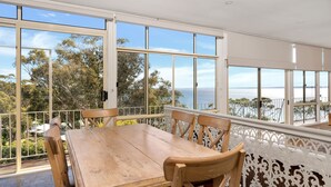 A dining area with a wooden table and chairs, large windows offering a view of the sea and trees. - Sea Point at Hyams Beach (Hyams Beach)