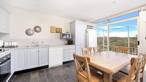 A modern kitchen with white cabinetry, a dining area with a wooden table and chairs, and a large window offering a scenic view. - Sea Point at Hyams Beach (Hyams Beach)
